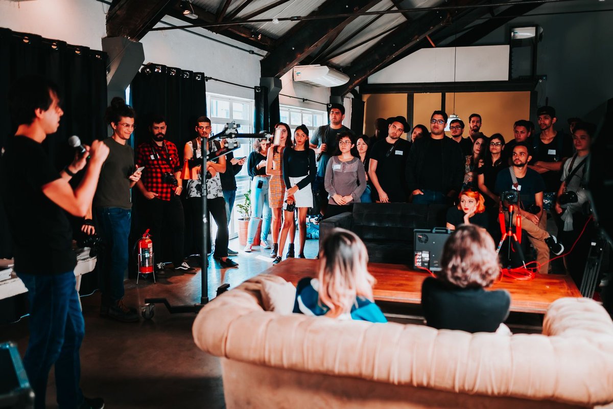 Speaker with a microphone briefing a standing production crew in an industrial studio with camera gear on a coffee table