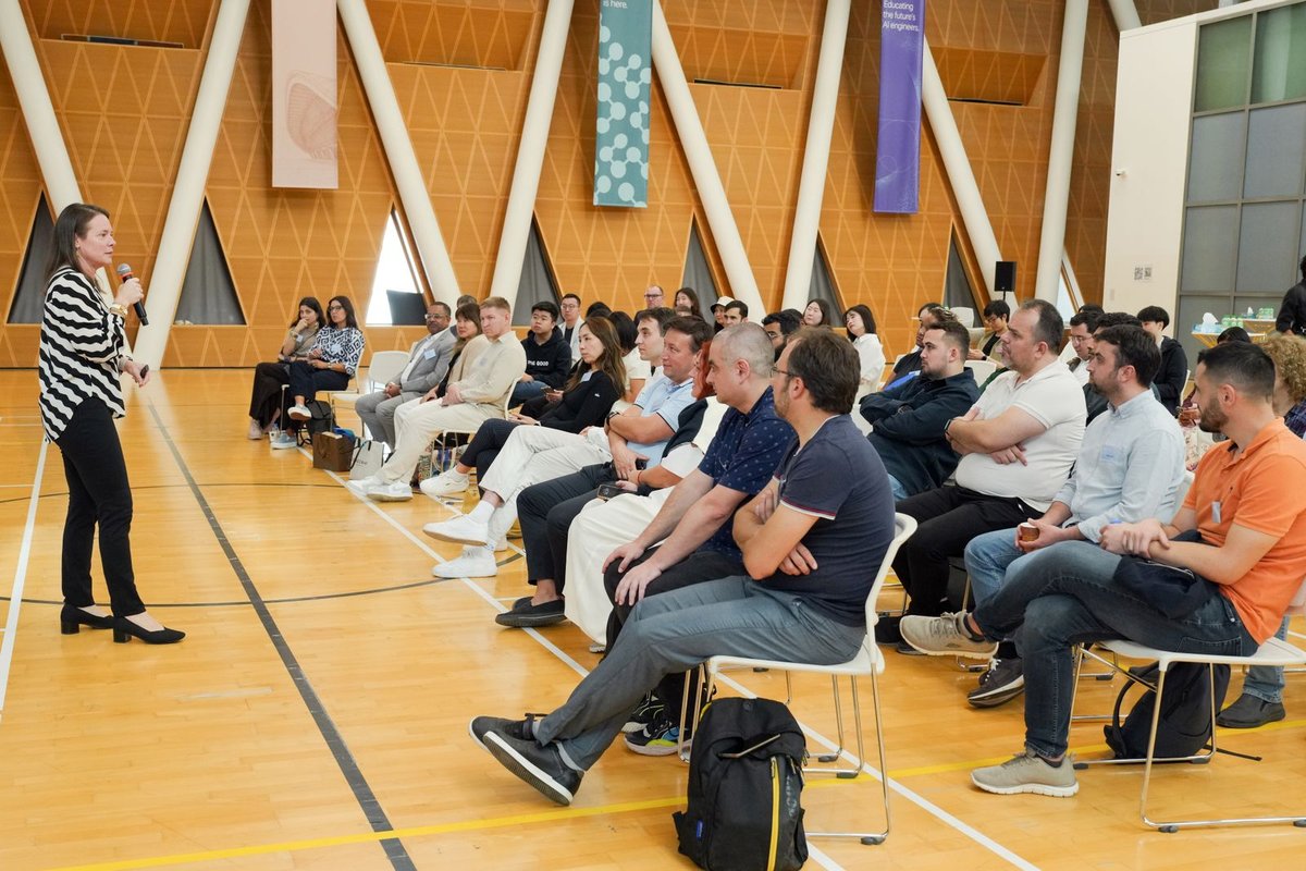 Woman with a microphone addressing rows of seated attendees in a bright multipurpose hall with wooden geometric walls